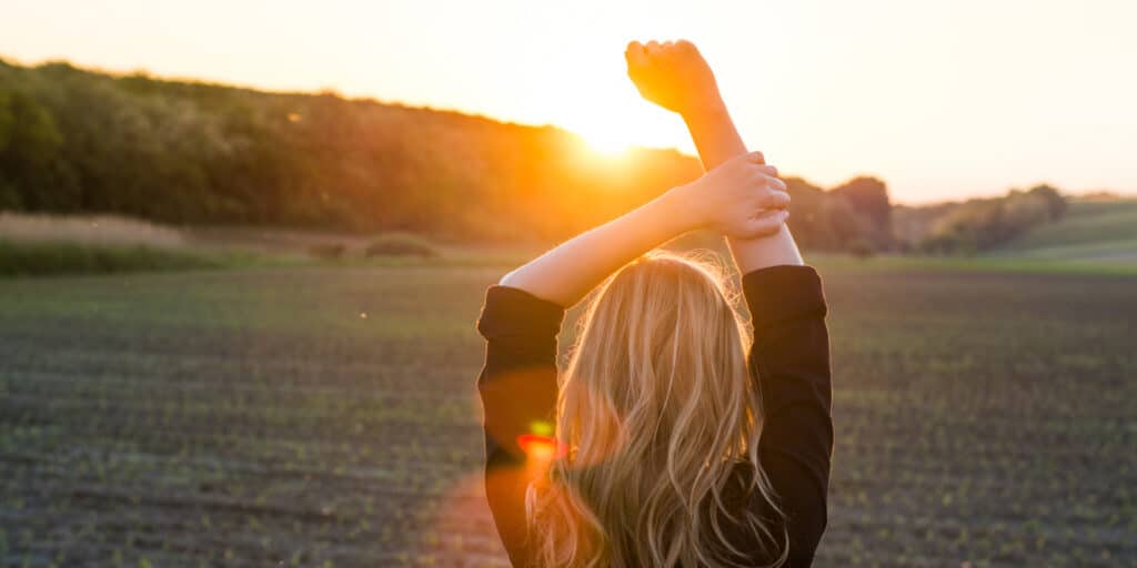 The back of a woman wearing a black long sleeved shirts that are rolled up holder her arms up with a field and the setting sun in the background.