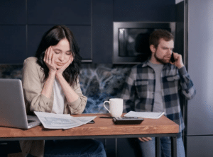 Woman looks stressed at desk with tax documents; man talks on phone in background.