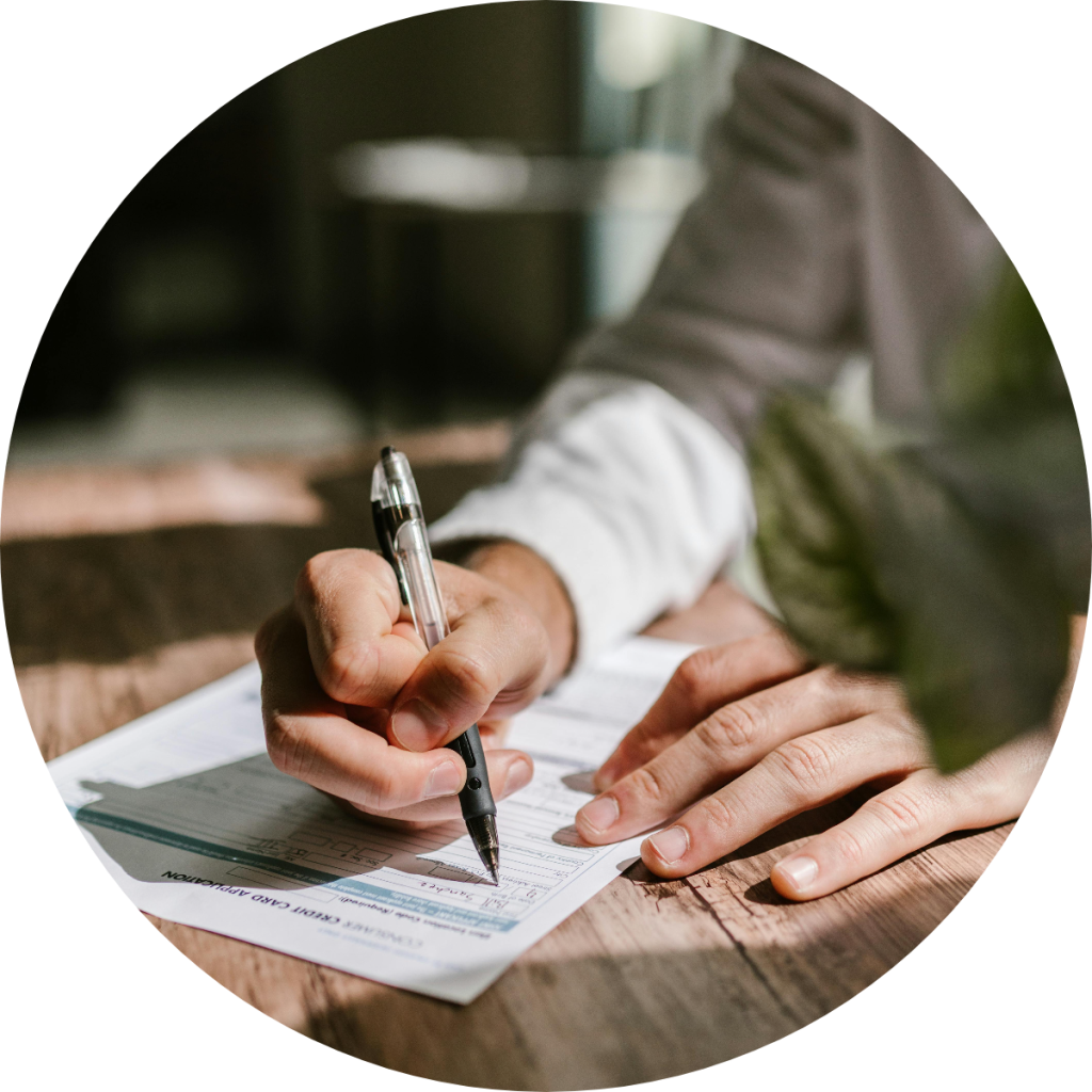 Person signing or filling out a credit card application form with pen on wooden desk during personal finance or loan application process
