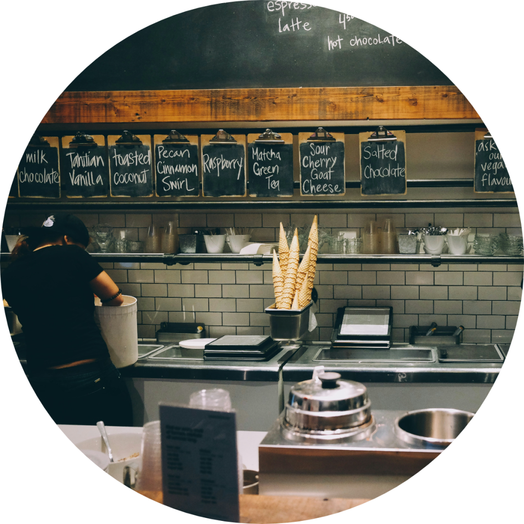 Artisan ice cream shop counter with chalkboard menu displaying flavors including Tahitian Vanilla, Matcha Green Tea, and Salted Chocolate with waffle cones and barista, small business retail concept