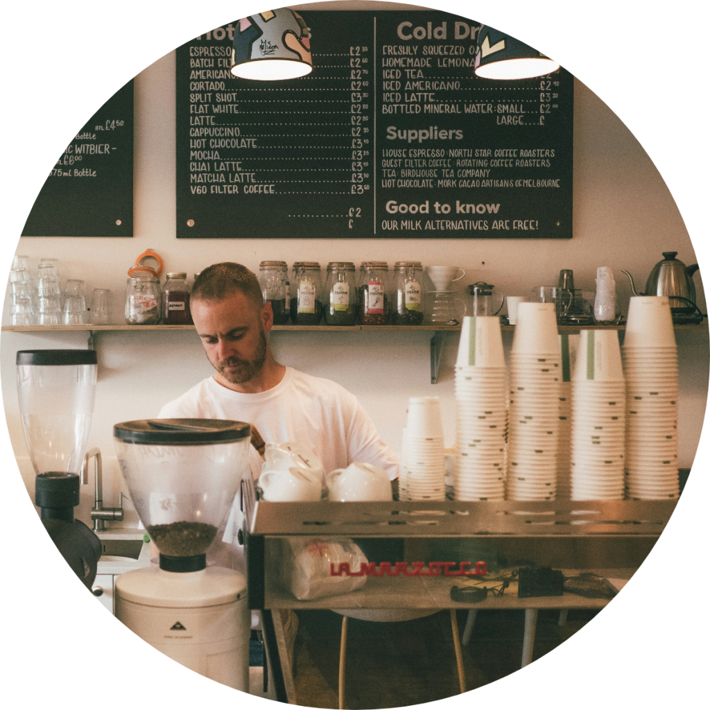 Male barista operating espresso machine at independent coffee shop with detailed hot and cold drinks chalkboard menu and stacked takeaway cups, small business food service concept